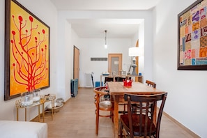 A dining area with a wooden table, chairs, and a red crown centerpiece. There is a large abstract painting on the left and a quilt on the right wall.