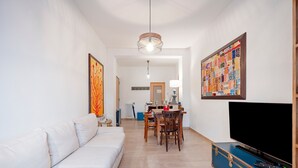 Un salon avec un canapé blanc, une table à manger avec des chaises, un téléviseur posé sur un meuble bleu et une œuvre d’art colorée accrochée au mur.