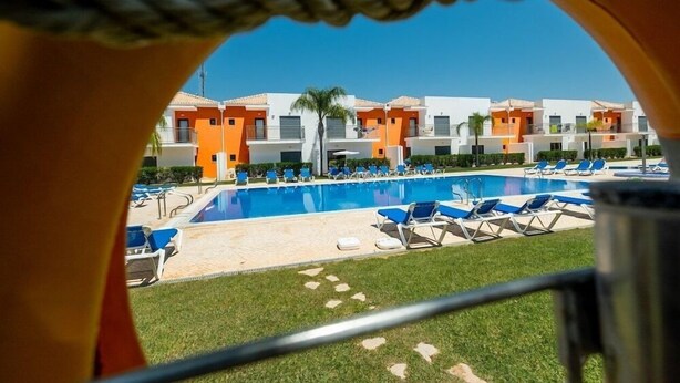 A swimming pool with blue lounge chairs, surrounded by a building with orange and white facades. - Jardins de Pera (Silves)
