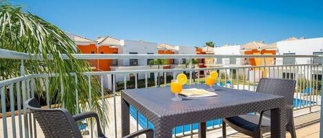A balcony with wicker furniture, a table with drinks, and a pool in the background.