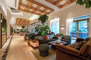 Un'ampia hall d'albergo con soffitto alto, travi in legno e una grande pianta da interno. Sono presenti diverse zone relax con divani e poltrone, e sullo sfondo si vede una reception.