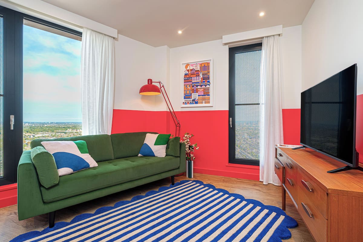 A modern living room with a green sofa, a red and white accent wall, a large window with a view, a flat-screen TV, and a blue-striped rug.