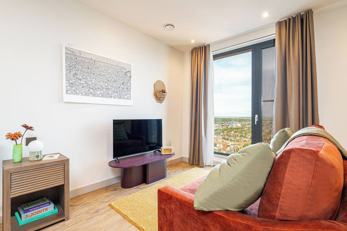 A modern living room with a flat-screen TV, a sofa, a small side table, and a large window with curtains.
