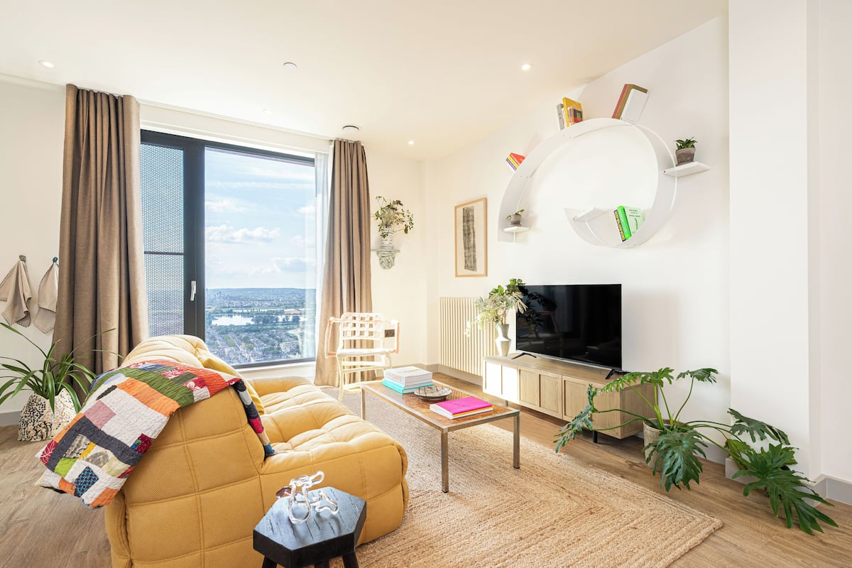 A modern living room with a sofa, coffee table, and TV. There is a large window with curtains, a plant, and a view of the city.