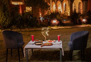 Une table à manger en plein air, avec des chaises, une pizza et des boissons, le tout installé devant un jardin avec un bâtiment en arrière-plan.