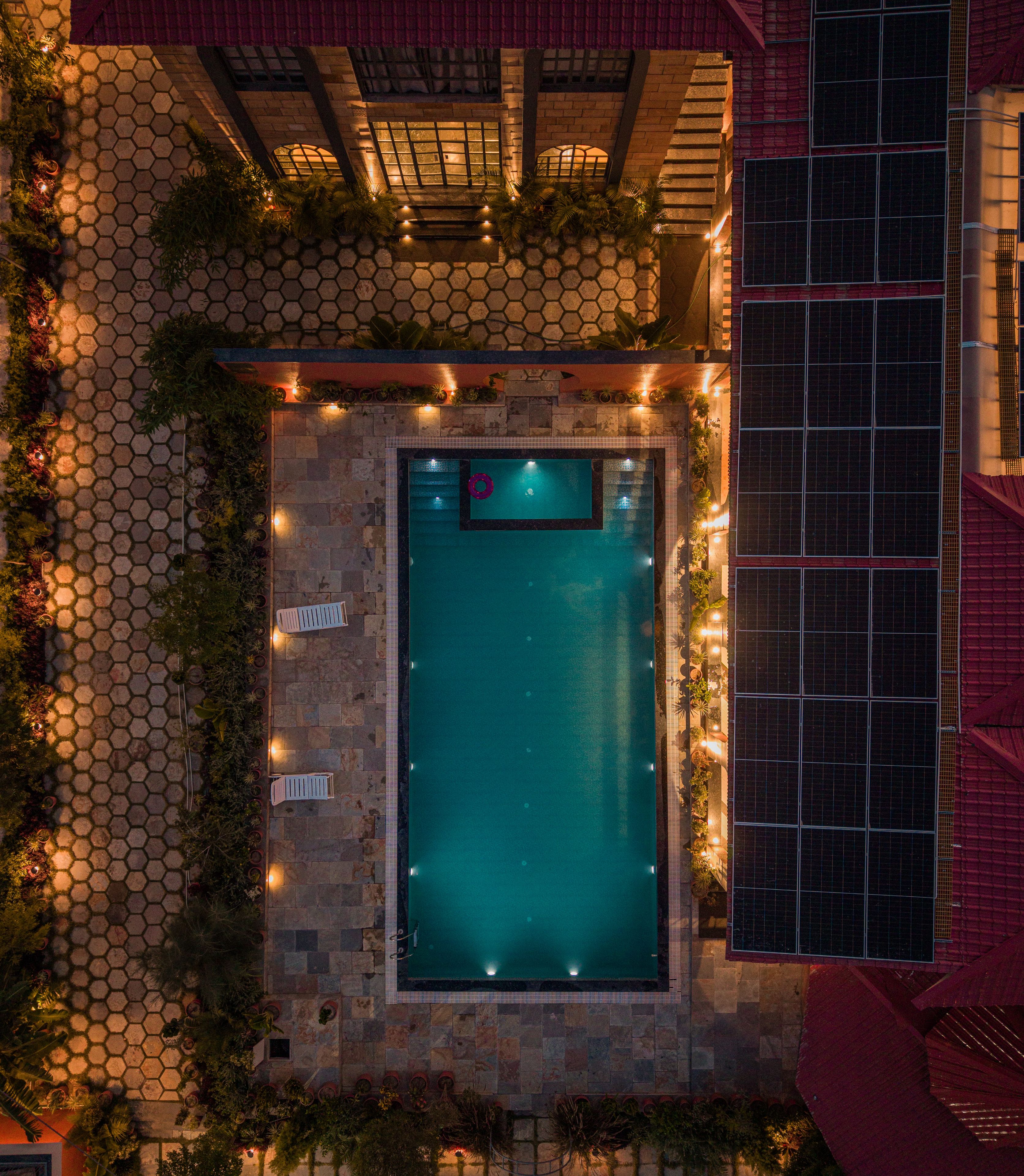 An aerial view of a swimming pool surrounded by hexagonal tiles, with a red-roofed building and solar panels to the right.