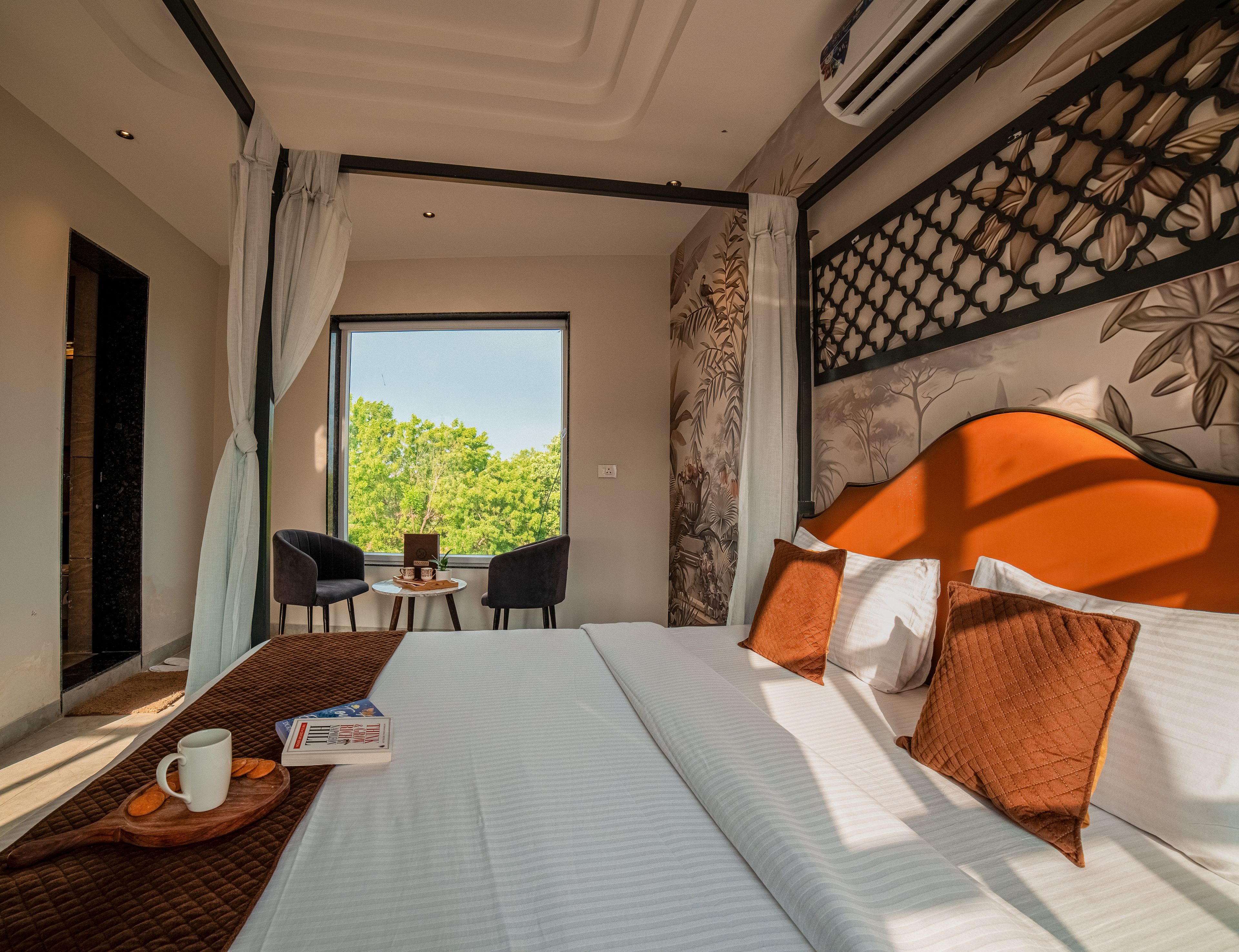 A hotel room with a large bed, a dining table with chairs, a window with a view of trees, and an air conditioning unit.