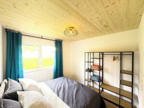 A bedroom with a bed, a window with curtains, a wooden ceiling, and a metal shelf unit. - Vacation home LieblingsKOOG in Friedrichskoog (Friedrichskoog)