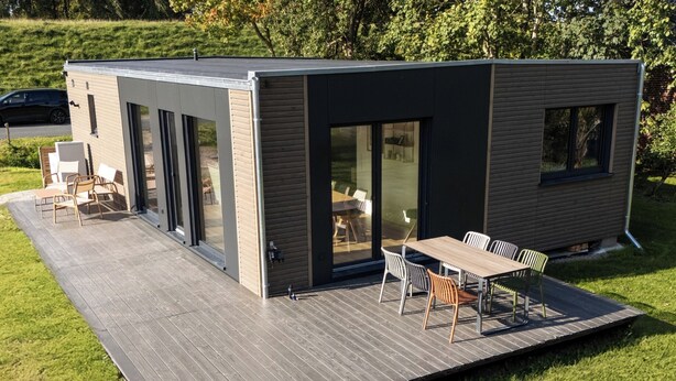 A modern, compact house with a flat roof, large sliding glass doors, and a patio area with outdoor furniture.