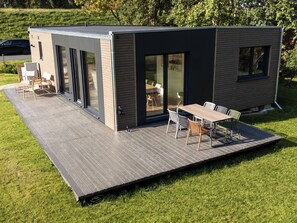 Une maison moderne et compacte, dotée d’un toit plat, de grandes portes-fenêtres coulissantes et d’un patio aménagé avec du mobilier d’extérieur.
