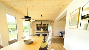 A modern kitchen with a wooden ceiling, a dining area with a long table and chairs, and a view of the outdoors through large windows.