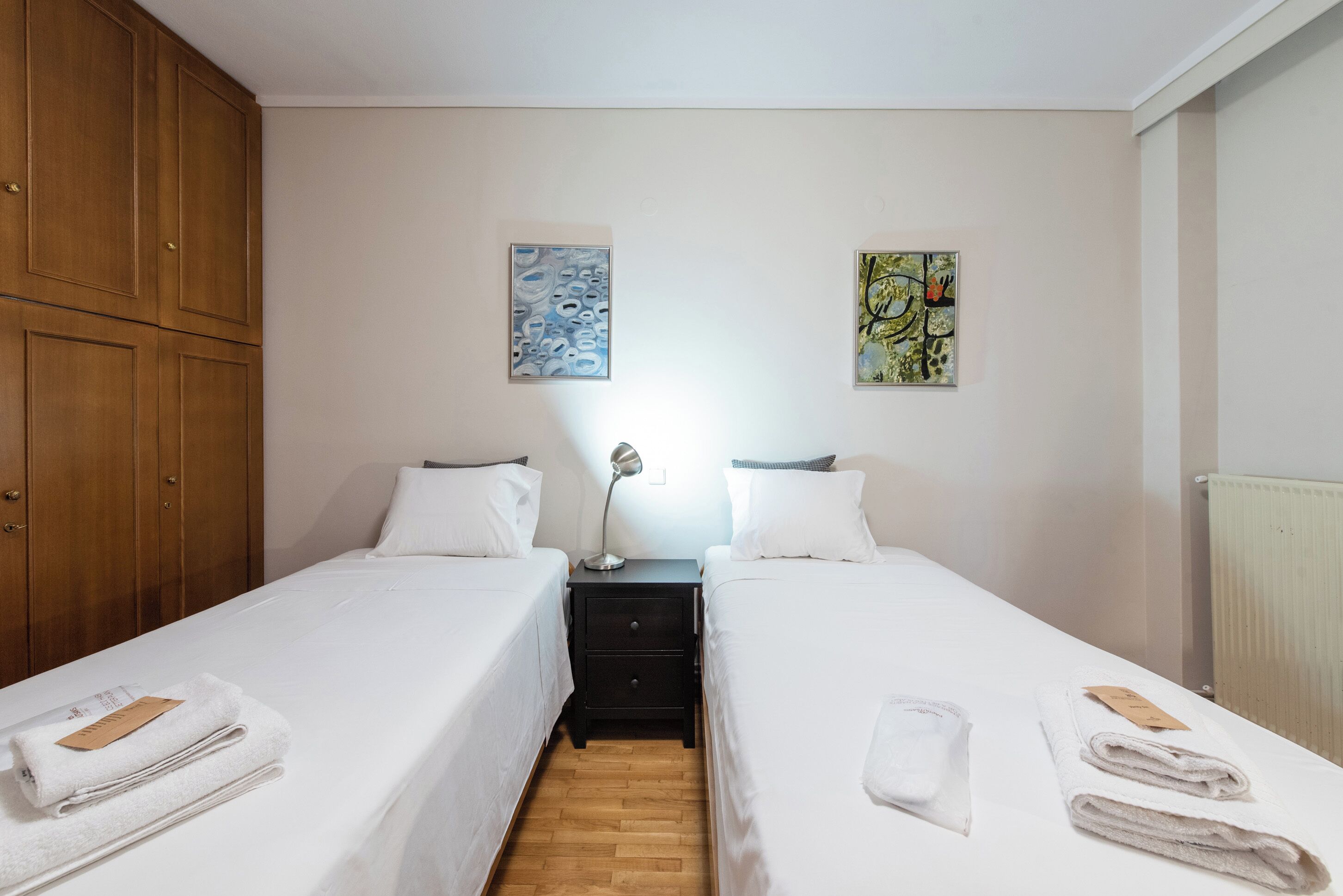 A hotel room with two single beds, a wooden wardrobe, a nightstand with a lamp, and artwork on the walls.