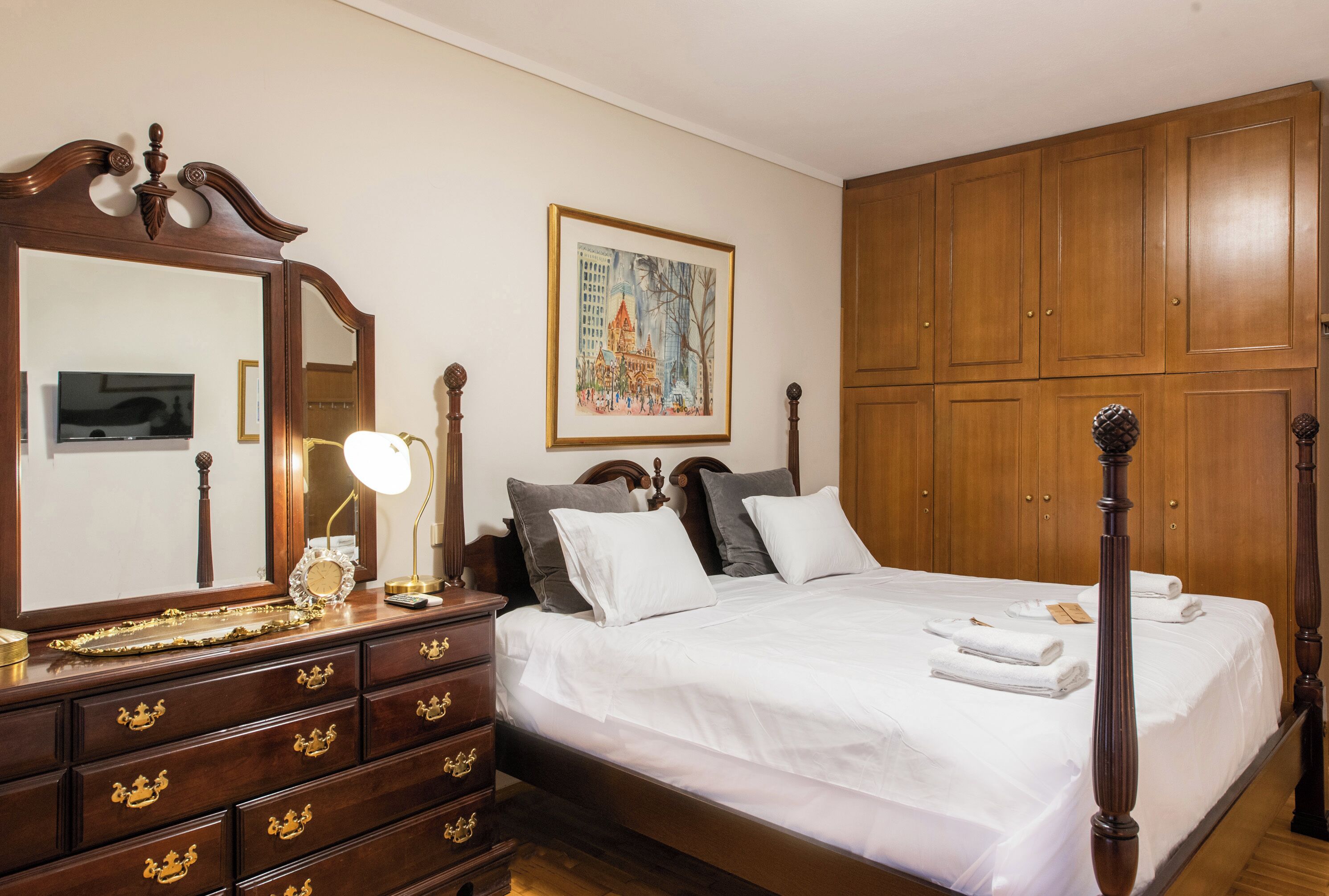 Una camera da letto ordinata con un letto grande, una cassettiera, uno specchio, un quadro incorniciato appeso al muro e mobili contenitori in legno.