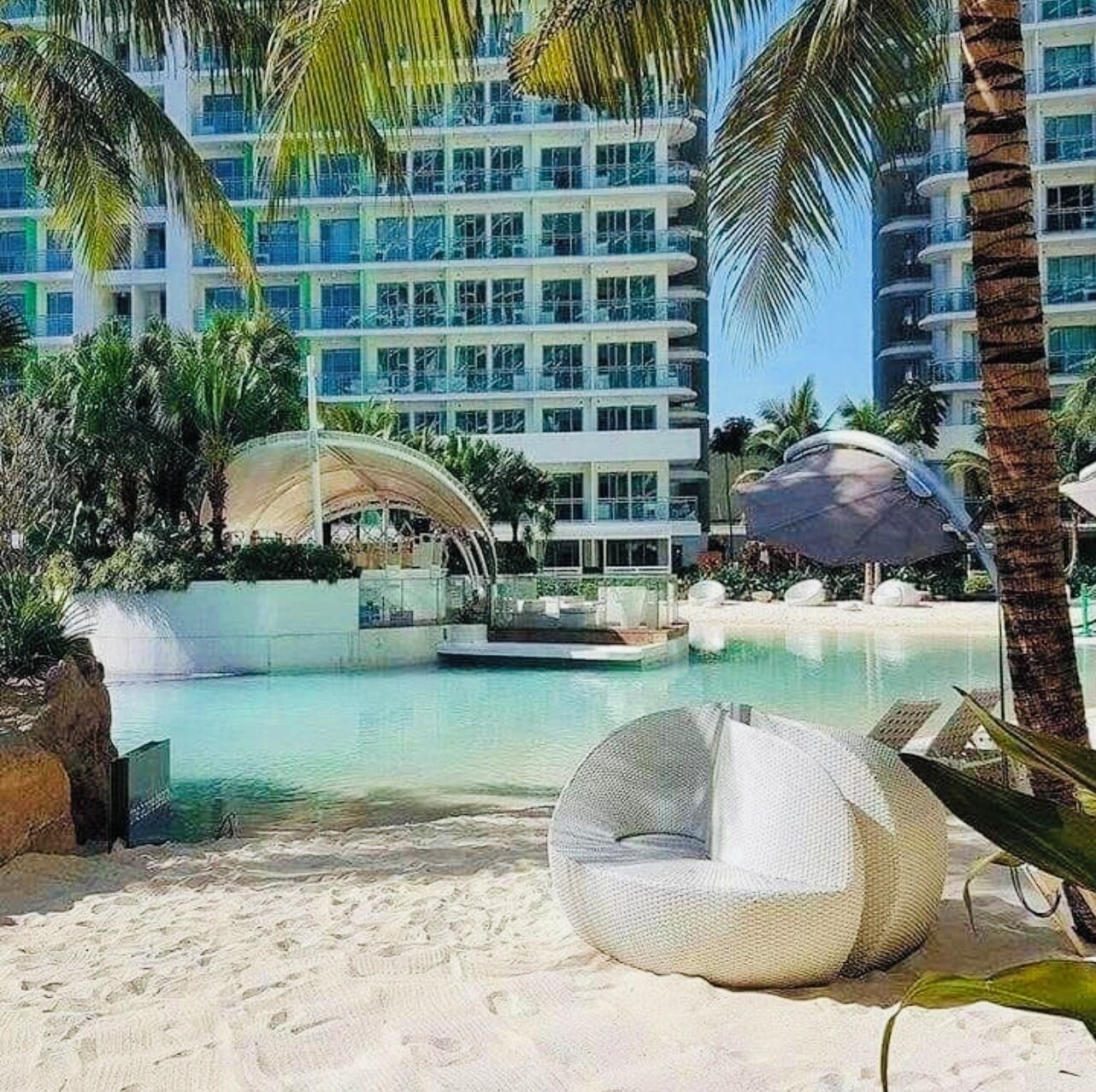 En pool vid ett semesterhotell med en sandstrand, solstolar och palmer.