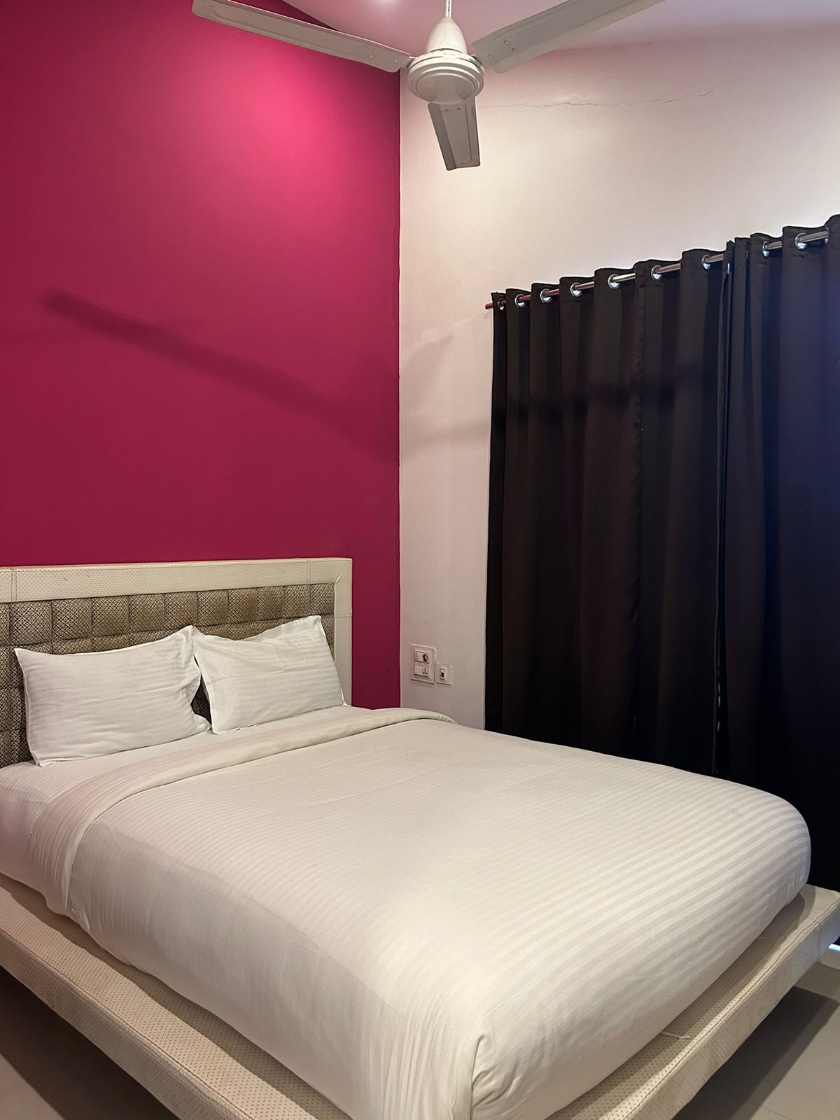 Una cama con ropa de cama blanca, un cabecero y un ventilador de techo en una habitación con paredes rosa y blancas.
