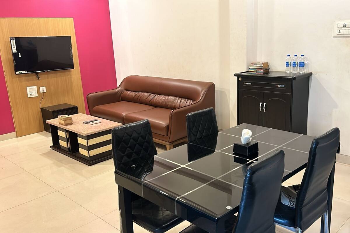 Habitación de hotel con un sofá de cuero marrón, una mesa de comedor negra con cuatro sillas, una pequeña mesita de madera, un mueble negro con un televisor montado en la pared y un dispensador de agua sobre el mueble.