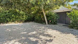 Uma entrada de cascalho leva a um edifício com porta escura, cercado por folhagem densa e um arbusto com flor vermelha.
