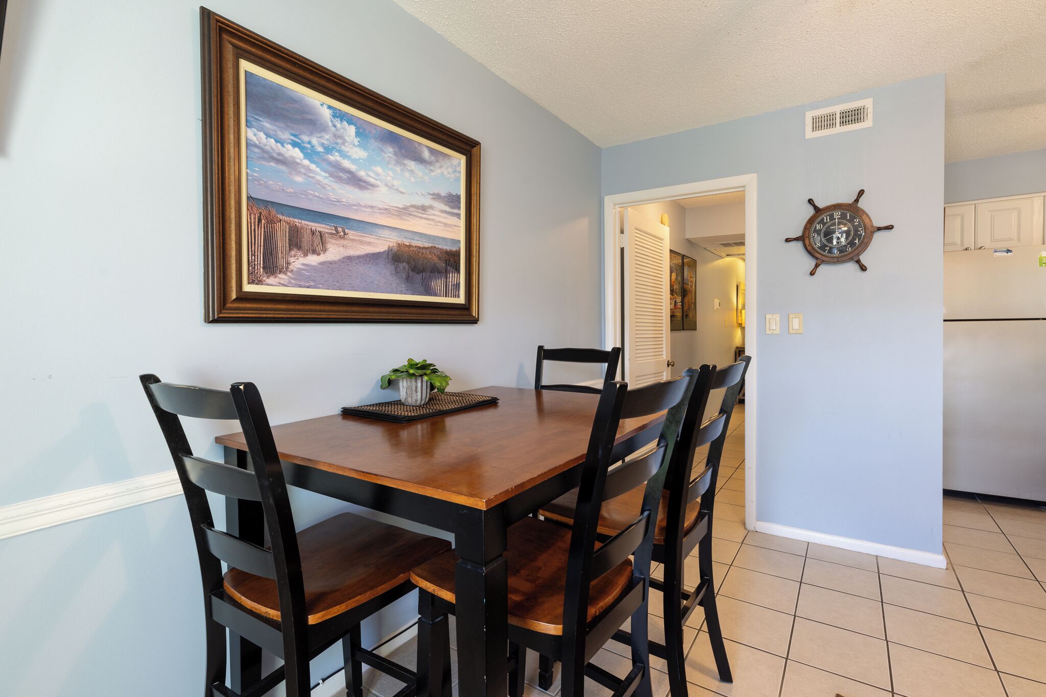 A dining area with a wooden table and chairs, a framed painting, and a ship's wheel on the wall.