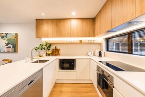A compact kitchen with wooden cabinets, a dishwasher, an oven, and a window with blinds. - Charming Home | Parking & Washer | Central City (Christchurch)