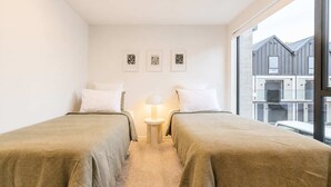 Habitación de hotel con dos camas, una mesita de noche, una lámpara y una ventana con vistas a edificios.