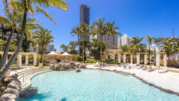 A resort pool with palm trees, lounge chairs, and a city skyline in the background. - Coastal Comfort 1BRM at Towers of Chevron (Gold Coast)