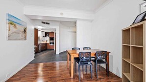 A dining area with a wooden table and chairs, a kitchen in the background, and a bookshelf to the right. - Coastal Comfort 1BRM at Towers of Chevron (Gold Coast)