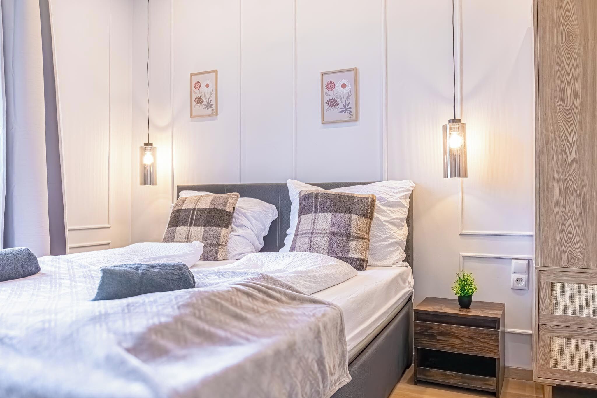 A neatly made bed with a checkered pillow, a bedside table with a plant, and wall-mounted lights.