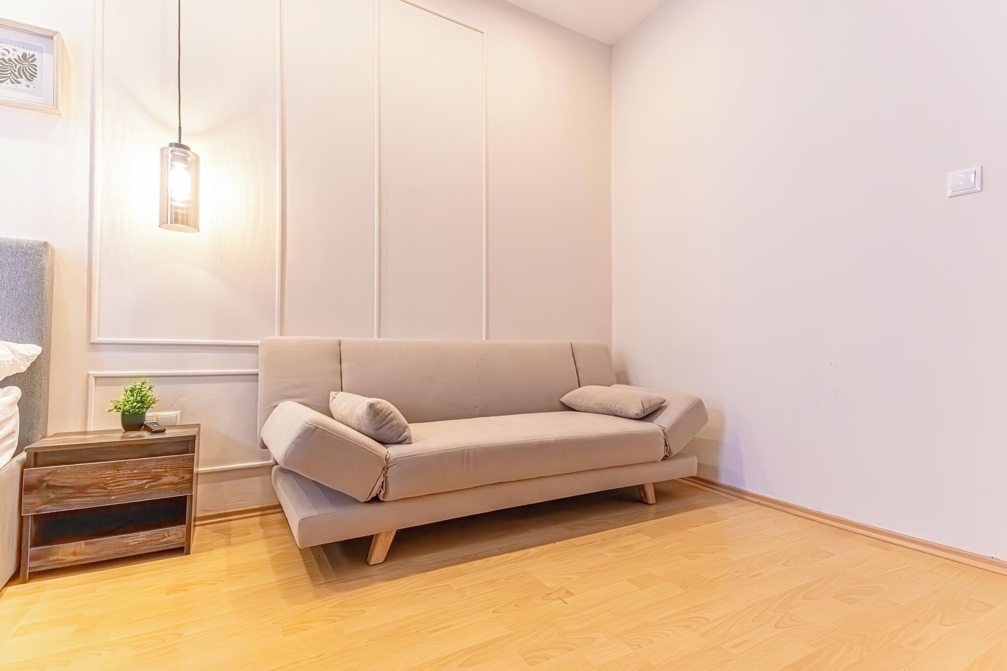 A minimalist living room with a beige sofa, a wooden side table with a plant, and a hanging light fixture.