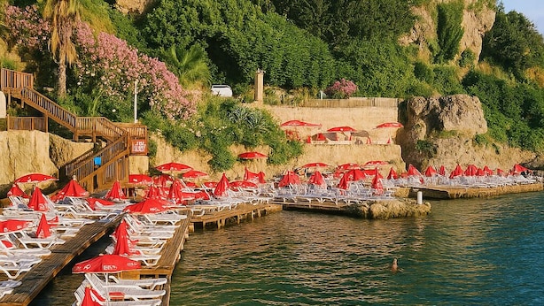 A coastal resort with wooden deck chairs and red umbrellas, a rocky cliff with greenery, and a clear blue sky. - Maxihomes Hadrian Nest (Antalya)