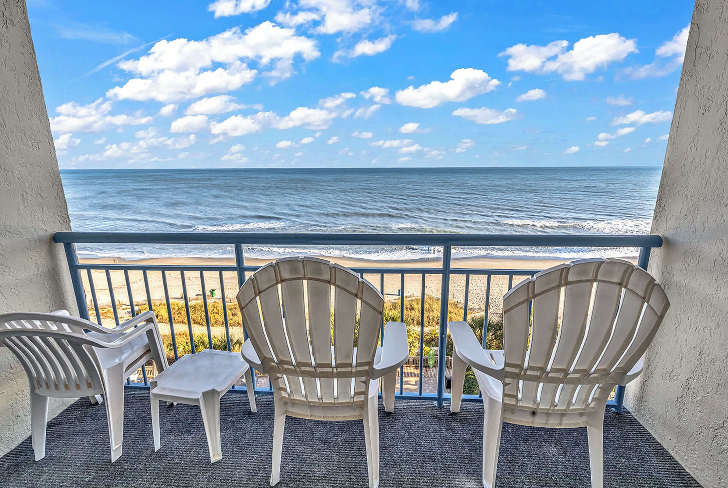Un balcon avec trois fauteuils Adirondack et une petite table, donnant sur une plage et l’océan.