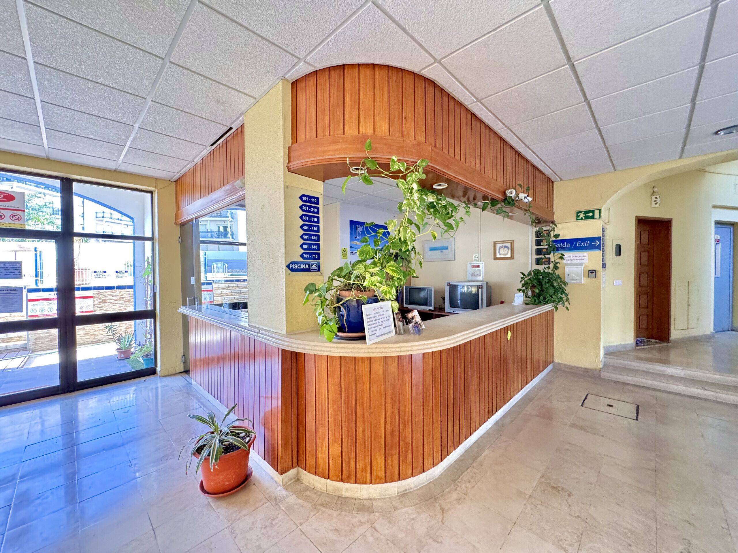 A reception desk with a wooden finish, a potted plant, and a sign indicating the location of the pool.