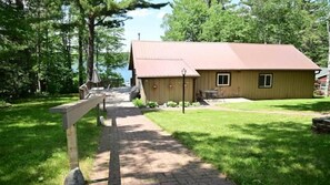 A wooden cabin with a sloped roof, a paved pathway leading to a wooden deck, and a grassy yard.