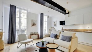 Un salon moderne avec un canapé, des fauteuils et une table basse. On y trouve également un coin cuisine avec des placards et un luminaire suspendu.