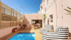 Un área junto a la piscina con muelle de madera, un sofá a rayas, una mesa con sillas y luces colgantes.