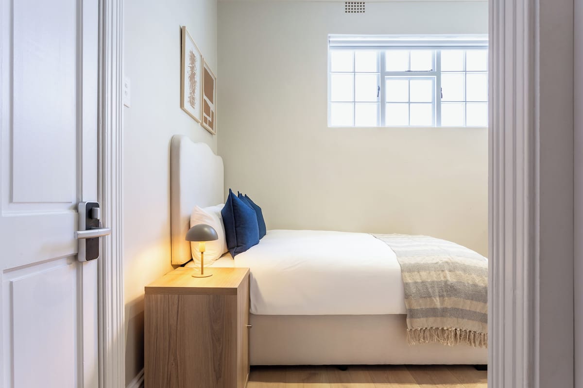 A bedroom with a bed, bedside table, lamp, and a window with blinds.