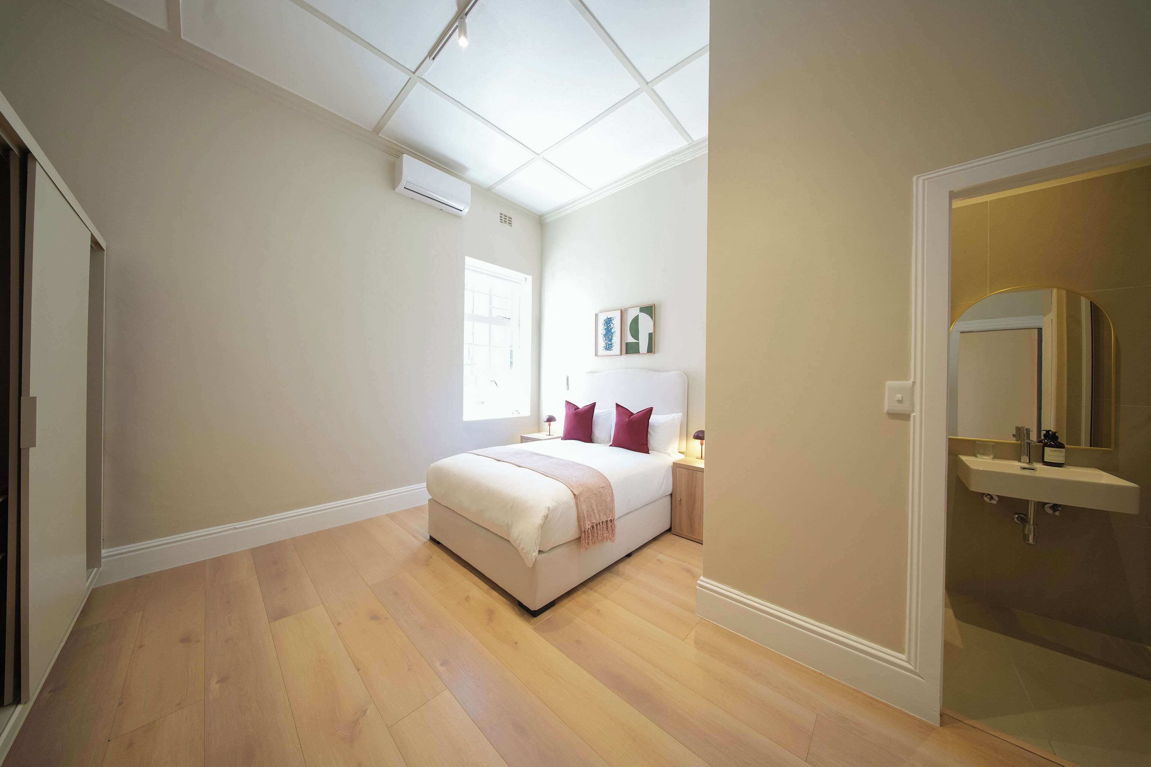 A bedroom with a bed, bedside tables, a window, and a bathroom with a sink and mirror.