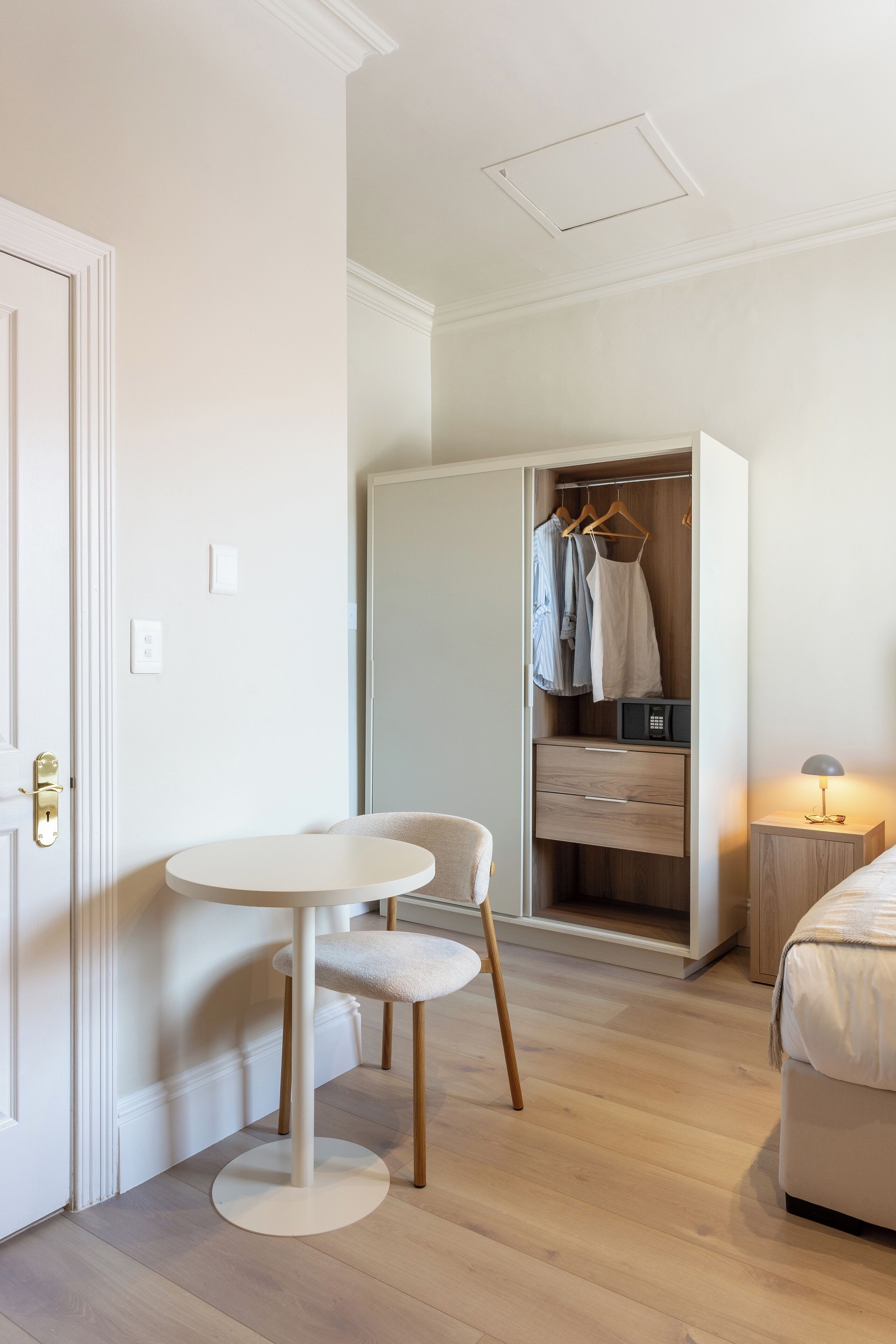 A room with a round table, a chair, a bed, a wardrobe, and a bedside lamp.