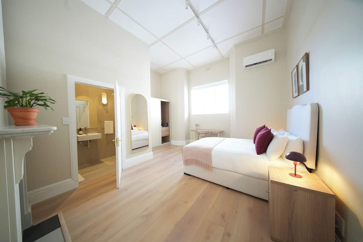 A bedroom with a bed, bedside table, and a nightstand. There is a window providing natural light and a door leading to another room.