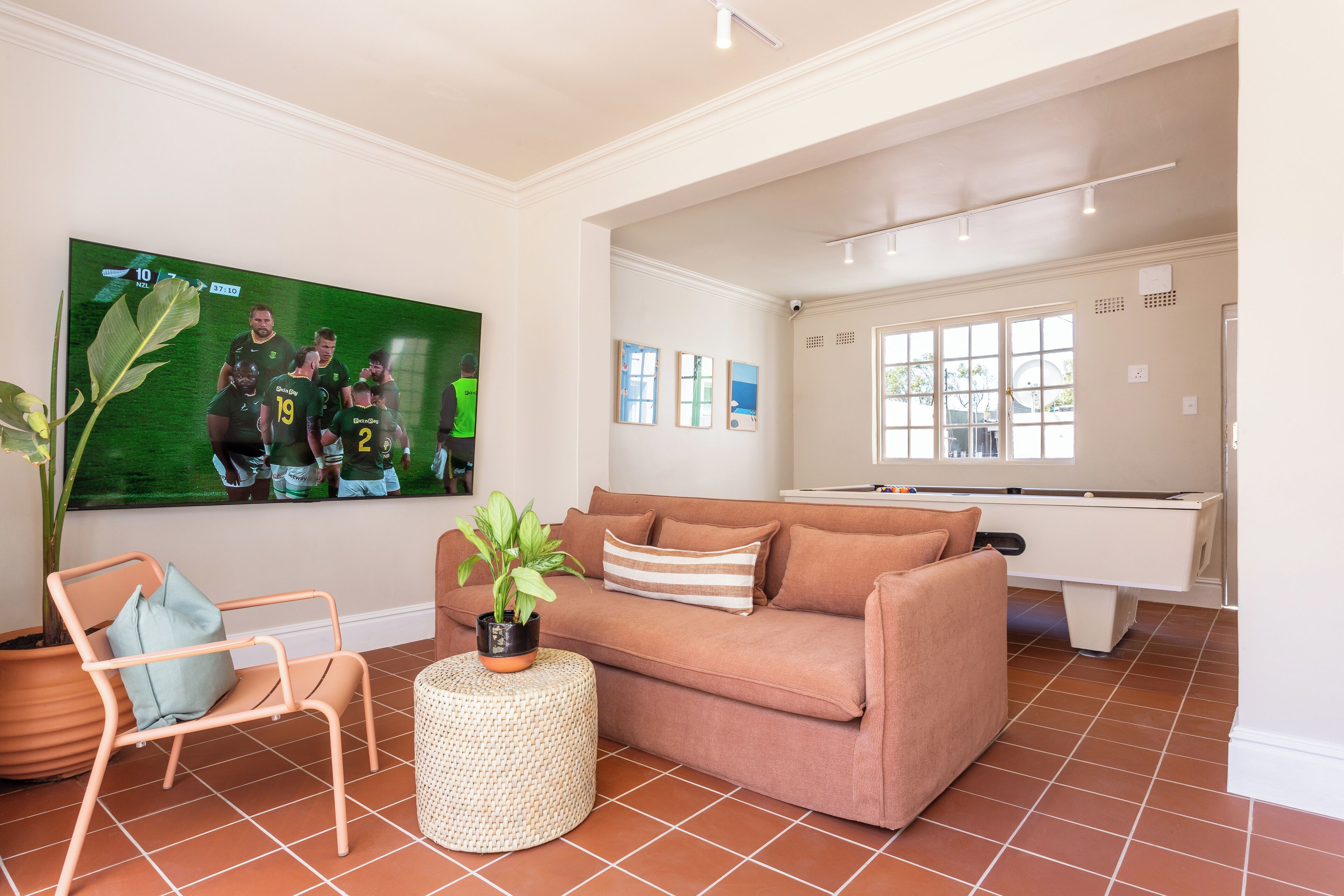 A living room with a sofa, armchair, potted plant, and a television displaying a sports event.