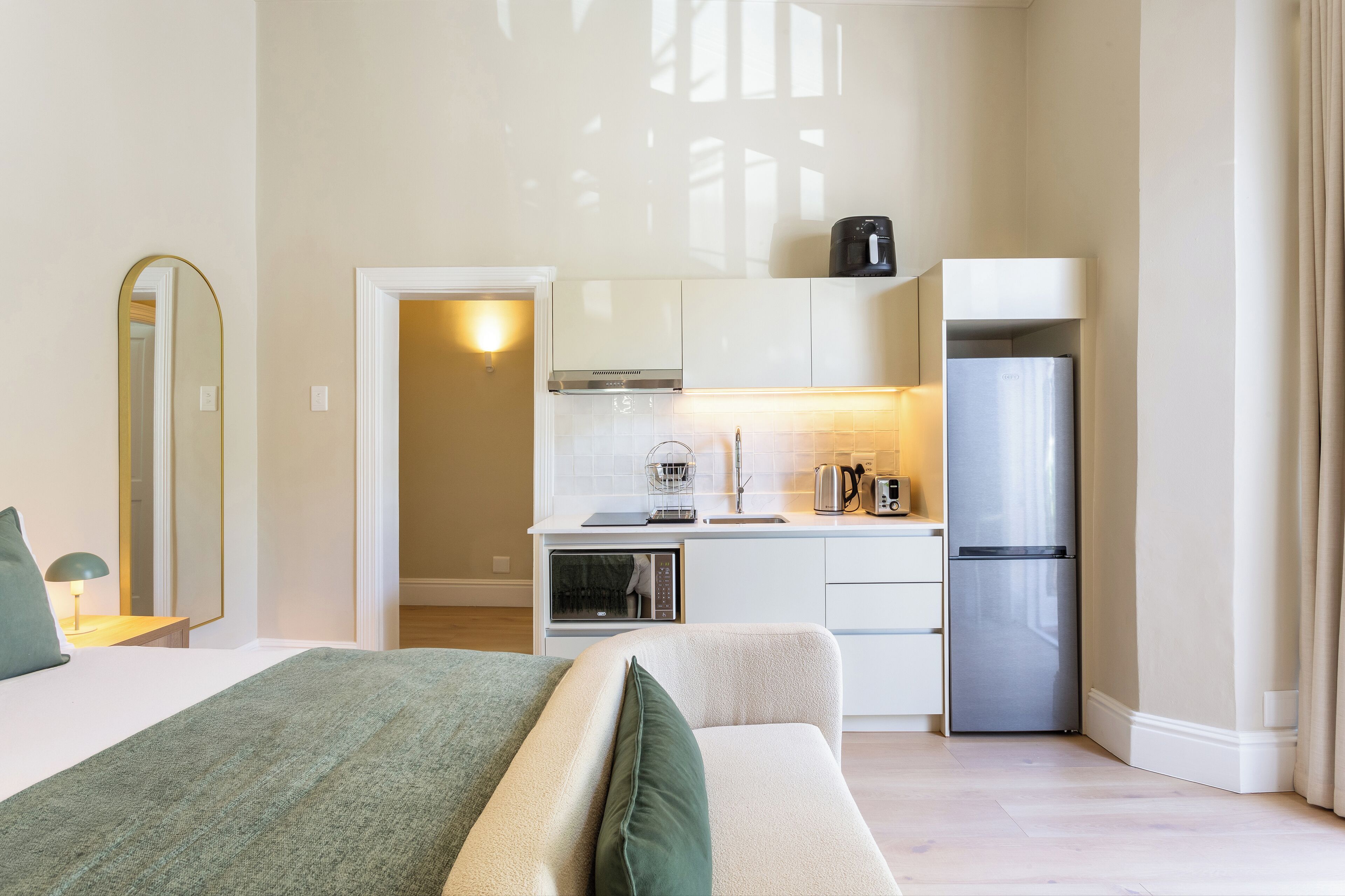 Una habitación de hotel moderna con una cama, kitchenette, un espejo y una puerta que da a otra habitación.