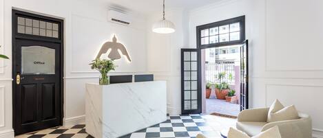 Uma área de recepção com balcão de mármore, uma porta preta com a placa 'Office', uma grande escultura iluminada e piso xadrez.