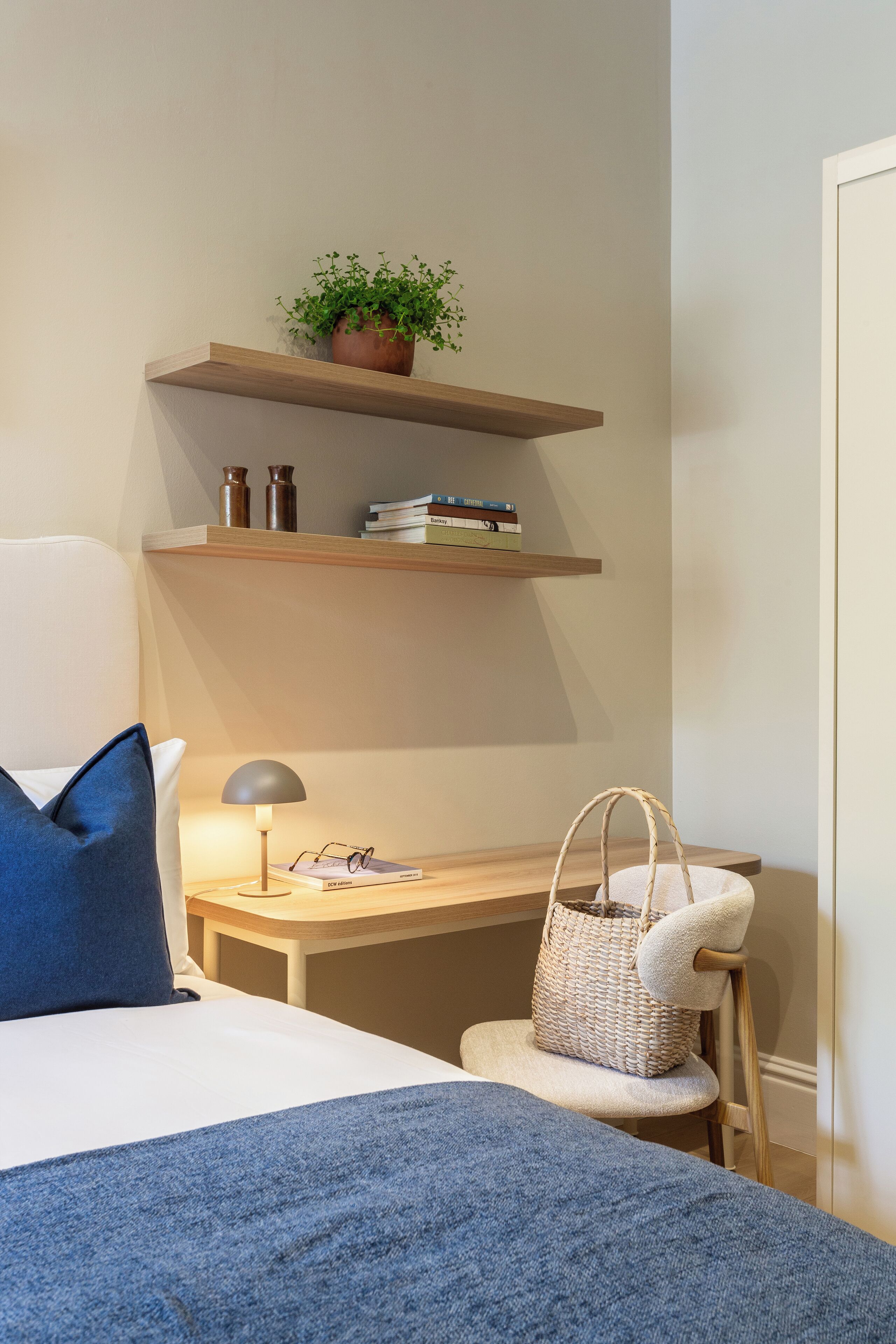 Une chambre à coucher comprenant un lit, un bureau, une chaise, une lampe, une étagère avec des livres et une plante, ainsi qu’une table de chevet avec un panier.