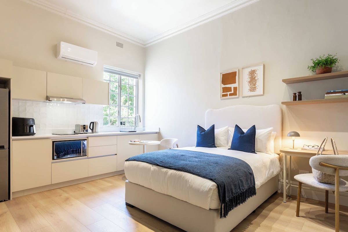 A modern hotel room with a kitchenette, a bed with blue pillows, a small table with a lamp, and a chair.