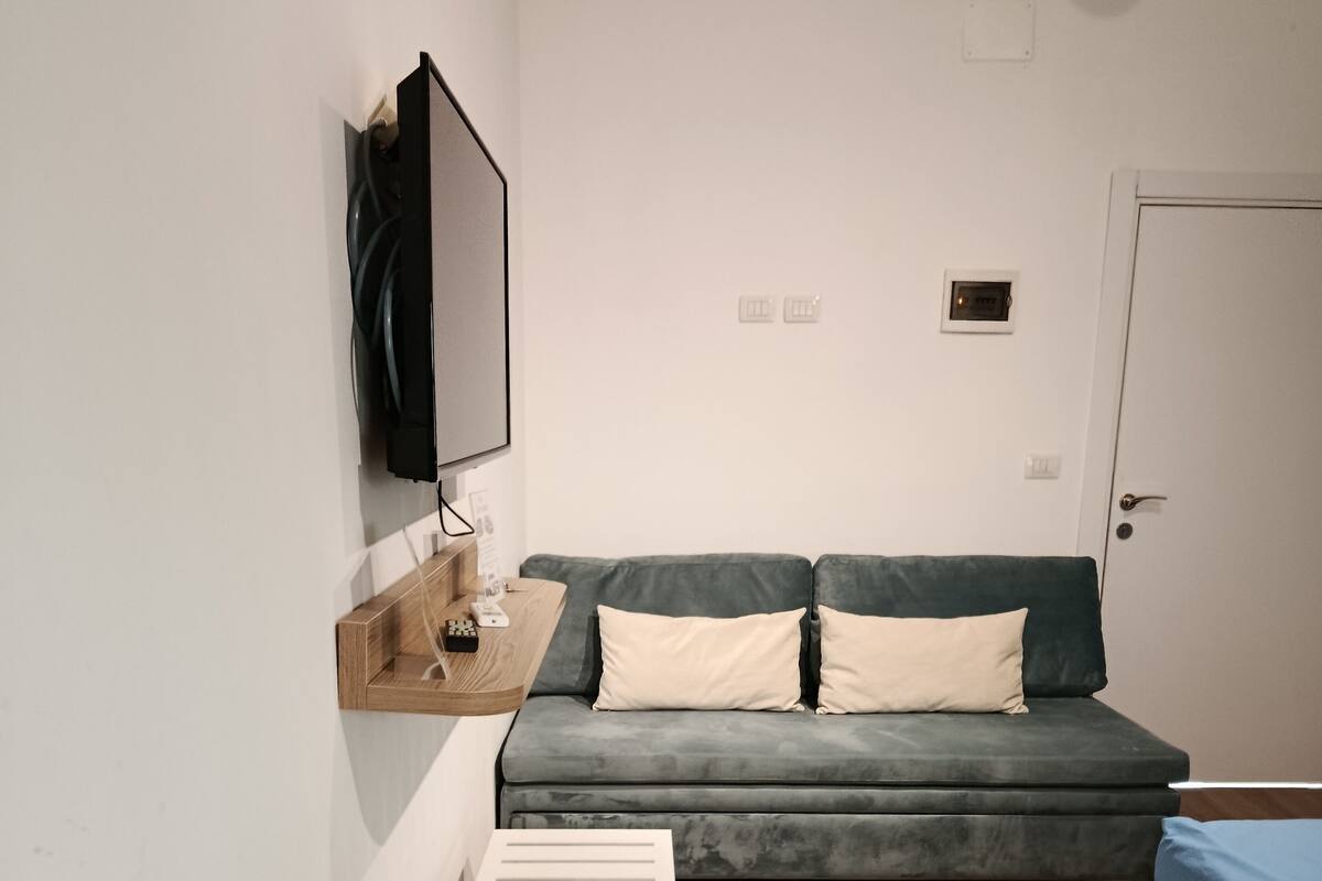 Une chambre moderne et minimaliste, avec un canapé gris, une petite table blanche et un lit recouvert d’une couverture bleue.