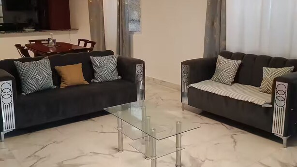 Ein Wohnzimmer mit zwei dunklen Sofas, einem Glastisch, einem Esstisch mit Stühlen und einem an der Wand befestigten Fernseher.