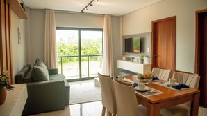 A modern living room with a sofa, dining table, and chairs. There is a TV mounted on the wall, a sliding glass door leading to a balcony, and a wooden cabinet. - Apto com 2 su tes a 100m do Japaratinga Resort (Japaratinga)