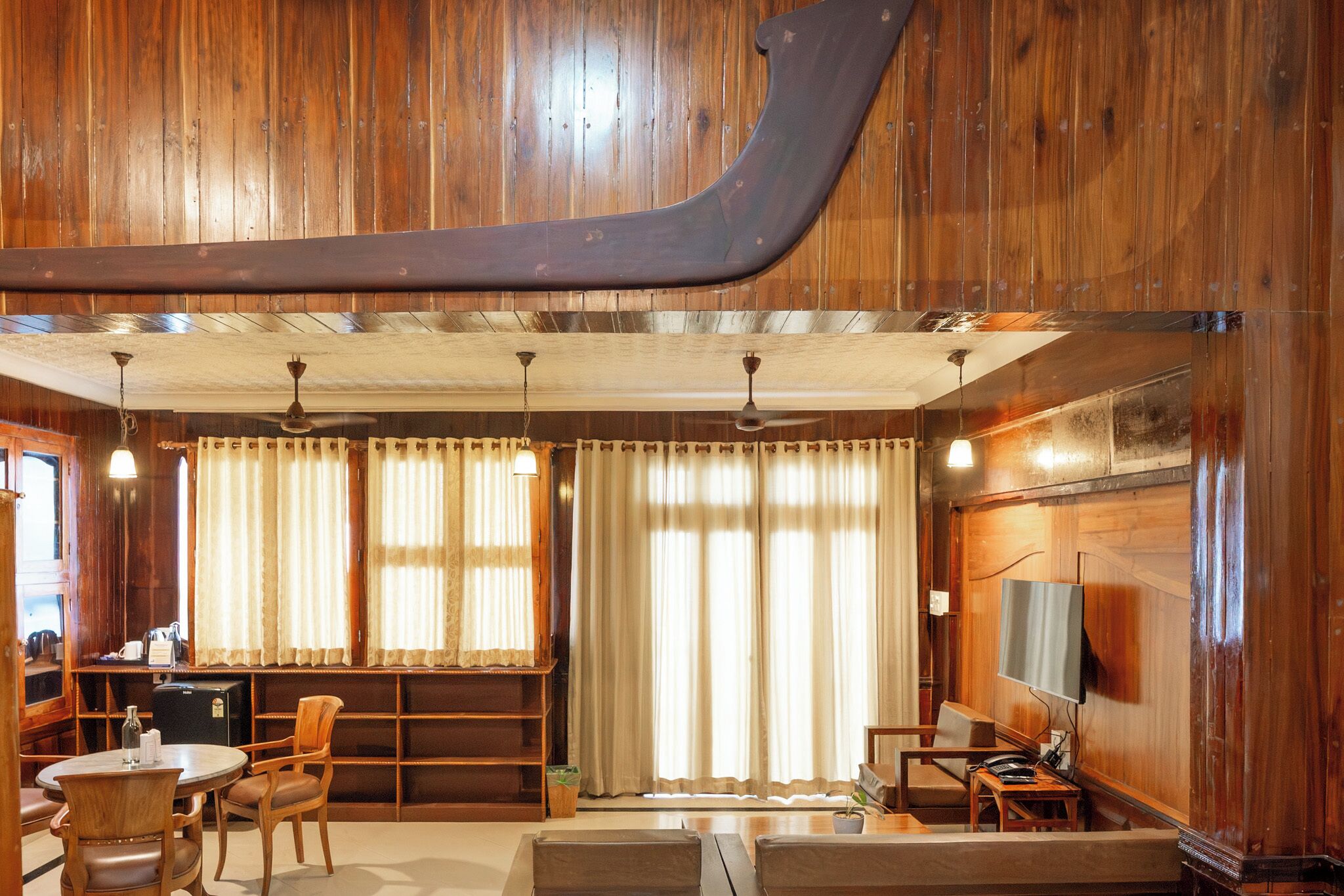 Una habitación con paneles de madera, una viga de madera grande, un ventilador de techo, un televisor y una zona de estar con sillas y una mesa.