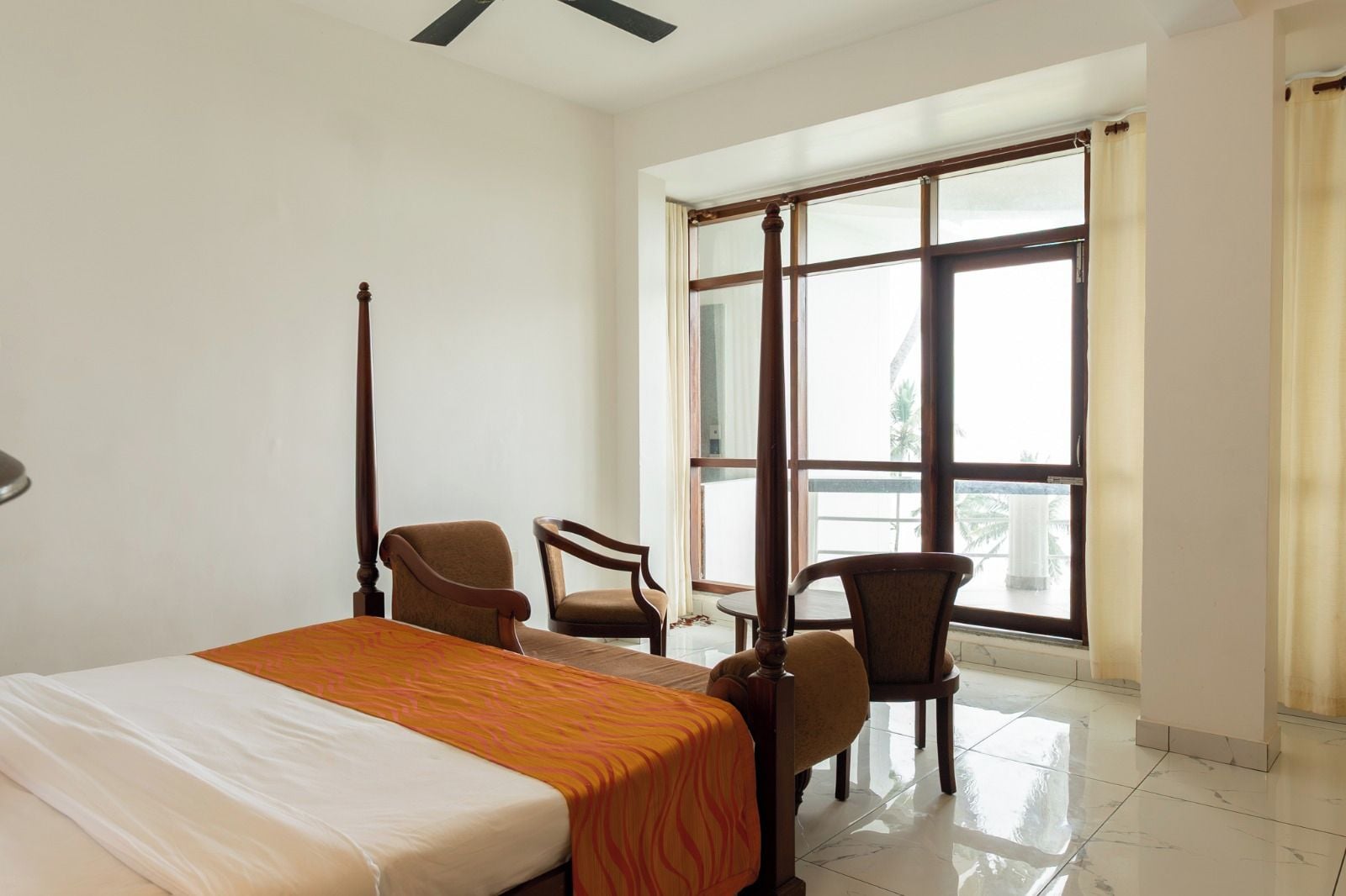 A hotel room with a bed, two chairs, a window with curtains, and a ceiling fan.