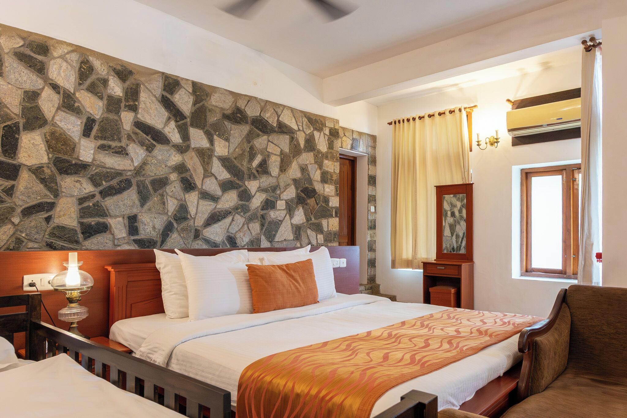Habitación de hotel con pared de piedra, cama con ropa blanca, cabecero de madera, mesita de noche con lámpara, silla de madera y ventana con cortinas.