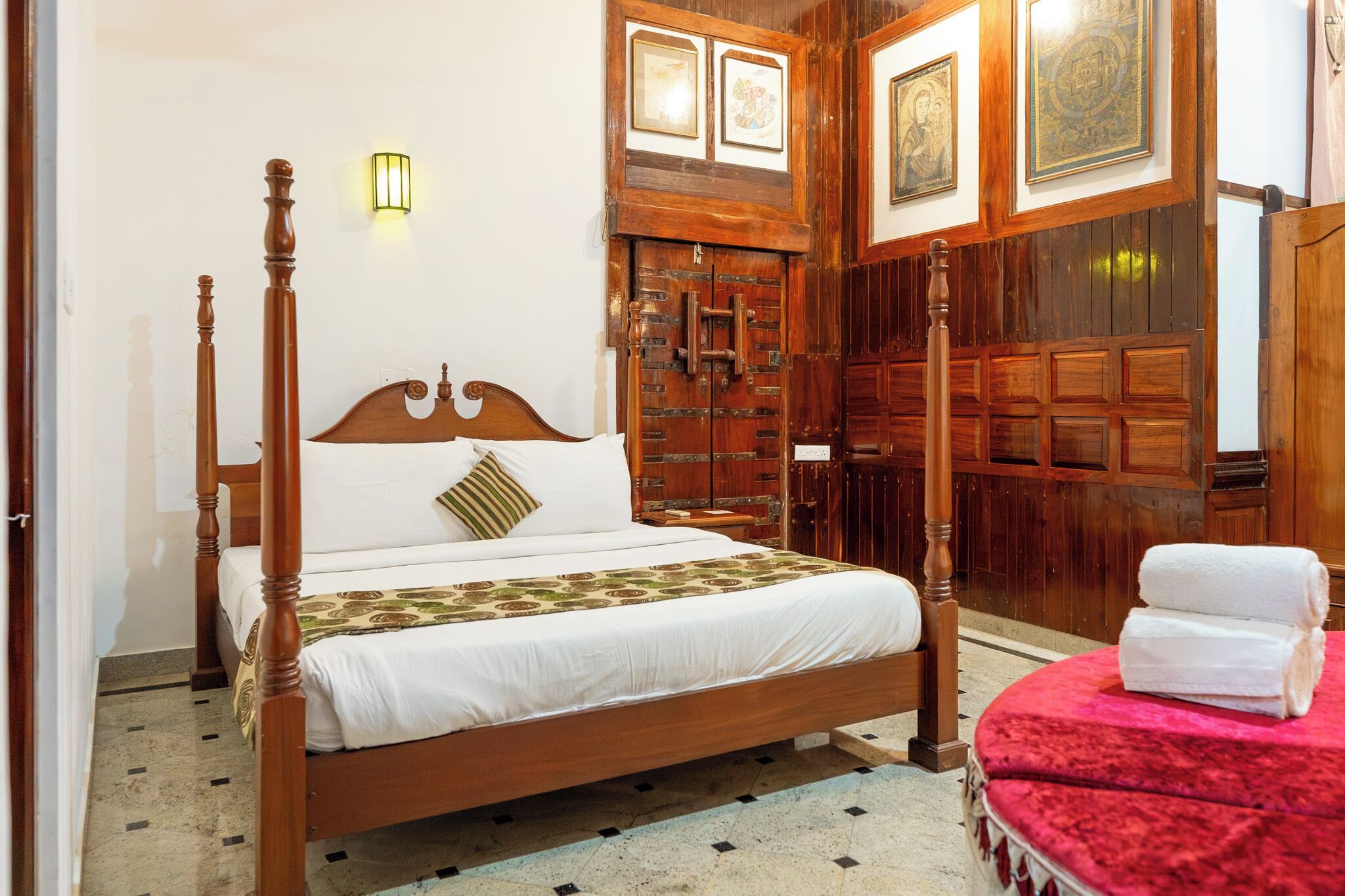Una cama con dosel y tablero de madera, una mesita de noche, un asiento con cojín rojo y una lámpara fijada a la pared.